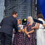 Na scenie kilkoro osób z nagrodami, dwie kobiety w czarno-różowych sukienkach. Tło ciemne, oświetlenie sceniczne.