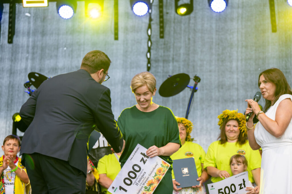 Na scenie kobieta odbiera nagrodę w wysokości 1000 zł w otoczeniu ludzi w jaskrawych koszulkach. Tło jest szare, a scena oświetlona żółtymi światłami. Obok stoi kobieta z mikrofonem.