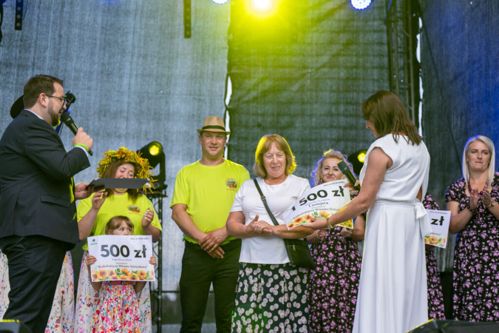 Na scenie grupa ludzi podczas wręczania nagrody. Kobieta z wieńcem trzyma tabliczkę z kwotą. Tło jest szare, światła żółte.