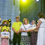 Na scenie grupa ludzi podczas wręczania nagrody. Kobieta z wieńcem trzyma tabliczkę z kwotą. Tło jest szare, światła żółte.