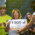 Na zdjęciu kobieta z nagrodą 1500 zł, z jasnym tłem. Obok niej dwie osoby, jedna w jasnej koszulce.