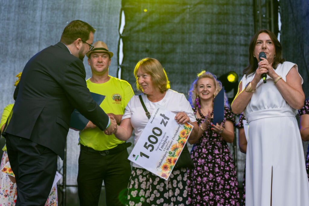 Na zdjęciu widać grupę ludzi na scenie. Kobieta trzyma dyplom z napisem 1500 zł. Tło jest ciemne, z jasnymi reflektorami.