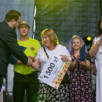 Na zdjęciu widać grupę ludzi na scenie. Kobieta trzyma dyplom z napisem 1500 zł. Tło jest ciemne, z jasnymi reflektorami.