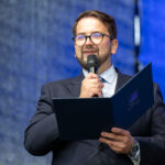 Mężczyzna w garniturze przemawia do mikrofonu, trzymając niebieską teczkę. Tło w odcieniach niebieskiego. Na twarzy ma okulary.