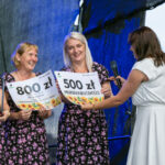 Cztery kobiety w kwiecistych sukienkach trzymają nagrody pieniężne. Tło ciemnoniebieskie. Jedna z kobiet ma mikrofon.