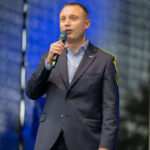 Mężczyzna w garniturze trzyma mikrofon. Tło jest ciemnoniebieskie, przypominając fale. Scena ma profesjonalny wygląd.