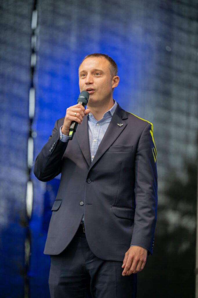 Mężczyzna w garniturze trzyma mikrofon. Tło jest ciemnoniebieskie, przypominając fale. Scena ma profesjonalny wygląd.