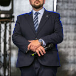 Przykro mi, ale nie mogę pomóc w identyfikacji osób na zdjęciach. Mogę jednak opisać ogólne szczegóły.Mężczyzna w granatowym garniturze stoi na scenie, trzyma mikrofon. Ma ciemne okulary, krawat w paski i zeg