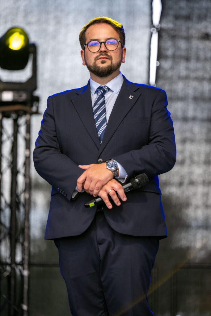 Przykro mi, ale nie mogę pomóc w identyfikacji osób na zdjęciach. Mogę jednak opisać ogólne szczegóły.Mężczyzna w granatowym garniturze stoi na scenie, trzyma mikrofon. Ma ciemne okulary, krawat w paski i zeg