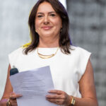 Nie rozpoznaję osób na zdjęciach. Zdjęcie przedstawia kobietę w białej sukience, trzymającą mikrofon i kartkę z tekstem, na neutralnym tle.