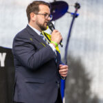 Mężczyzna w ciemnym garniturze z mikrofonem na scenie. W tle rozmazane światła i sprzęt muzyczny. Jasne, naturalne oświetlenie.