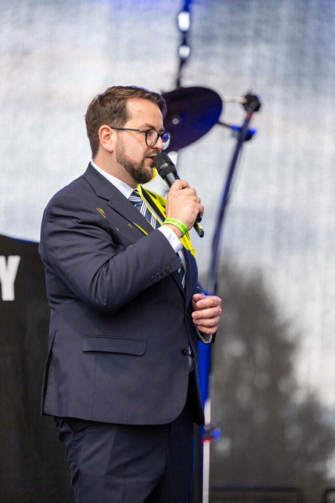 Mężczyzna w ciemnym garniturze z mikrofonem na scenie. W tle rozmazane światła i sprzęt muzyczny. Jasne, naturalne oświetlenie.