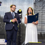 Na scenie stoi kobieta w białej sukni i mężczyzna w garniturze. Oboje trzymają mikrofony i mówią do publiczności. Tło jest ciemne z delikatnym oświetleniem.