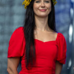 Kobieta w czerwonej sukience i wianku z żółtych kwiatów na tle ciemnego materiału. Ma długie czarne włosy. Uśmiecha się.