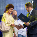 Wydarzenie na scenie. Kobieta w żółtej sukience odbiera dokument od mężczyzny w garniturze. Tło ciemne, kilku ludzi w tle.