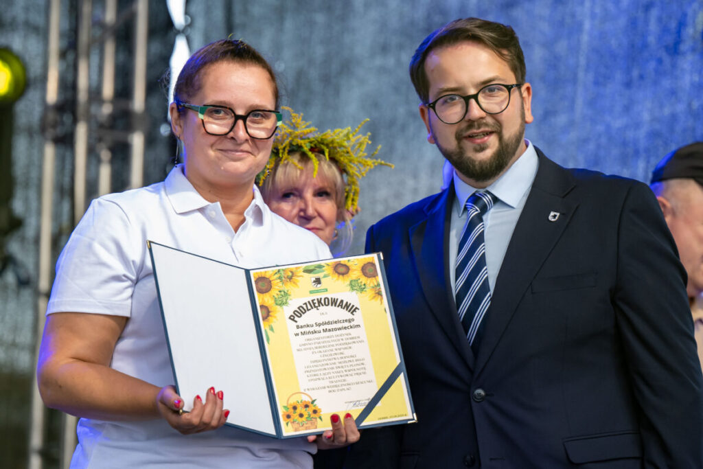 Na zdjęciu jest kobieta w białej koszuli i mężczyzna w garniturze, trzymają podziękowanie. Tło jest niebieskie. W tle widoczna jest osoba z wiankiem.