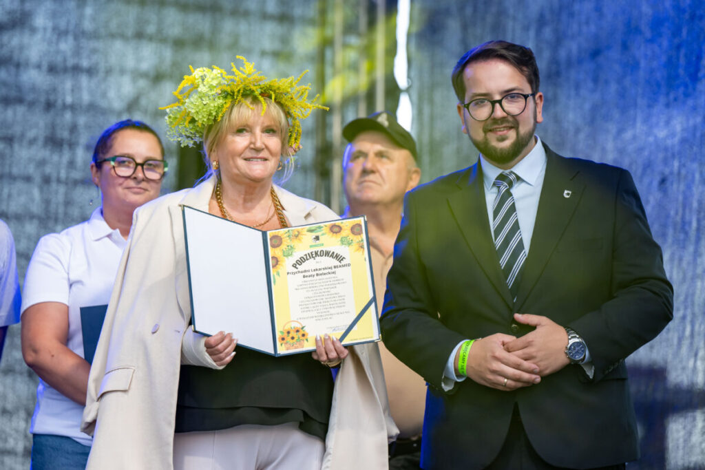 Grupka dzieci stoi na tle regałów bibliotecznych z książkami w reku trzymają zrobione przez siebie kolorowe palmy wielkanocne.