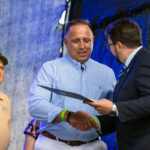 Trzech mężczyzn na scenie. Dwóch w garniturach uścisk dłoni, w tle starszy mężczyzna w czapce. Tło granatowe.