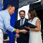 Trzy osoby w formalnych ubraniach na scenie, dwie z nich podają sobie ręce. Tło jest ciemne z niebieskimi akcentami. Kobieta trzyma niebieską teczkę.