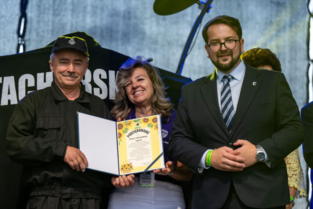 Na zdjęciu są trzy osoby, uśmiechnięte, trzymające dyplom w okładce. Tło sceniczne jest ciemne, dominują czernie. Mężczyzna w garniturze stoi po prawej.