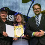 Na zdjęciu są trzy osoby, uśmiechnięte, trzymające dyplom w okładce. Tło sceniczne jest ciemne, dominują czernie. Mężczyzna w garniturze stoi po prawej.