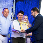 Na zdjęciu grupa osób, jedna z nich wręcza certyfikat. Tło w odcieniach niebieskiego. Oni są elegancko ubrani.