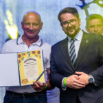 Na zdjęciu dwóch mężczyzn, jeden trzyma dyplom z żółtymi kwiatami. Tło jest niebiesko-szare, zdelikatnym światłem. Mężczyzna w garniturze nosi okulary.