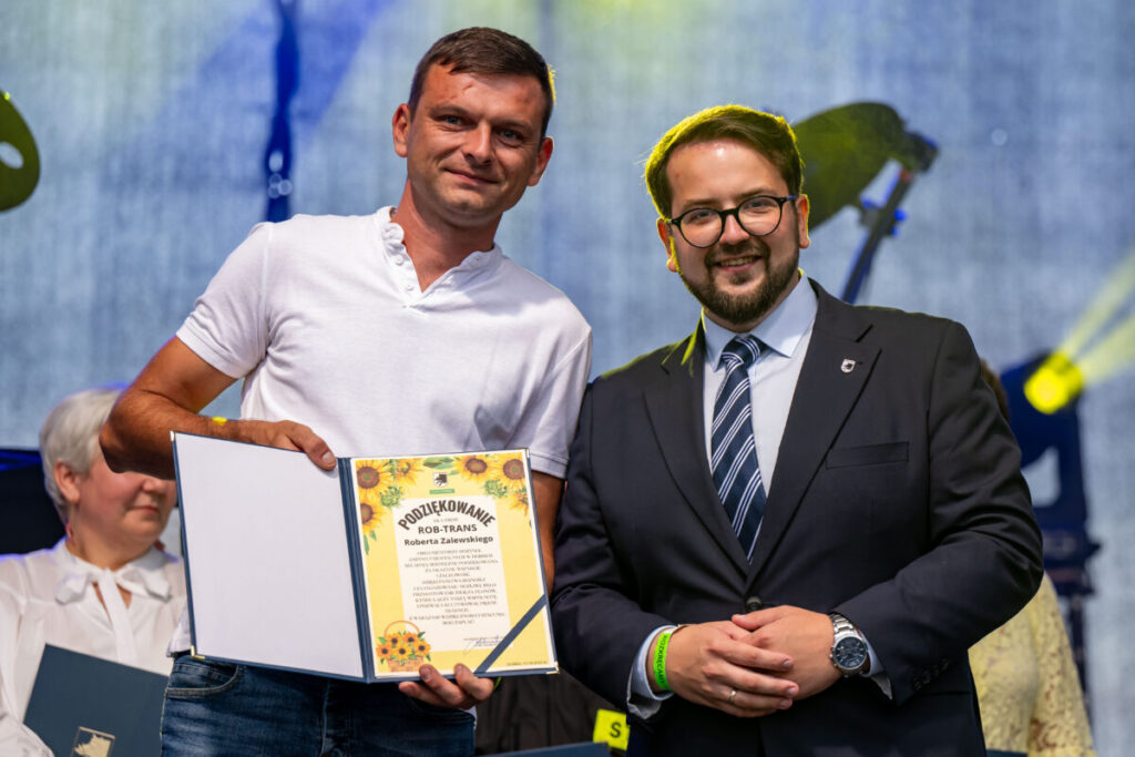 Dwóch mężczyzn na scenie; jeden w białej koszulce, trzyma certyfikat z motywem słoneczników. Drugi w garniturze, tło niebieskie.