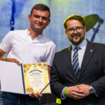 Dwóch mężczyzn na scenie; jeden w białej koszulce, trzyma certyfikat z motywem słoneczników. Drugi w garniturze, tło niebieskie.