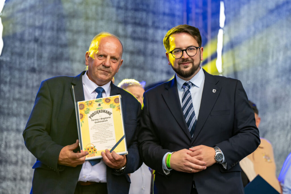Nie rozpoznaję osób na zdjęciach. Dwie osoby w garniturach na tle sceny. Jeden trzyma dyplom z napisem „Podziękowanie”. Tło jest ciemne, oświetlone żółtym światłem.