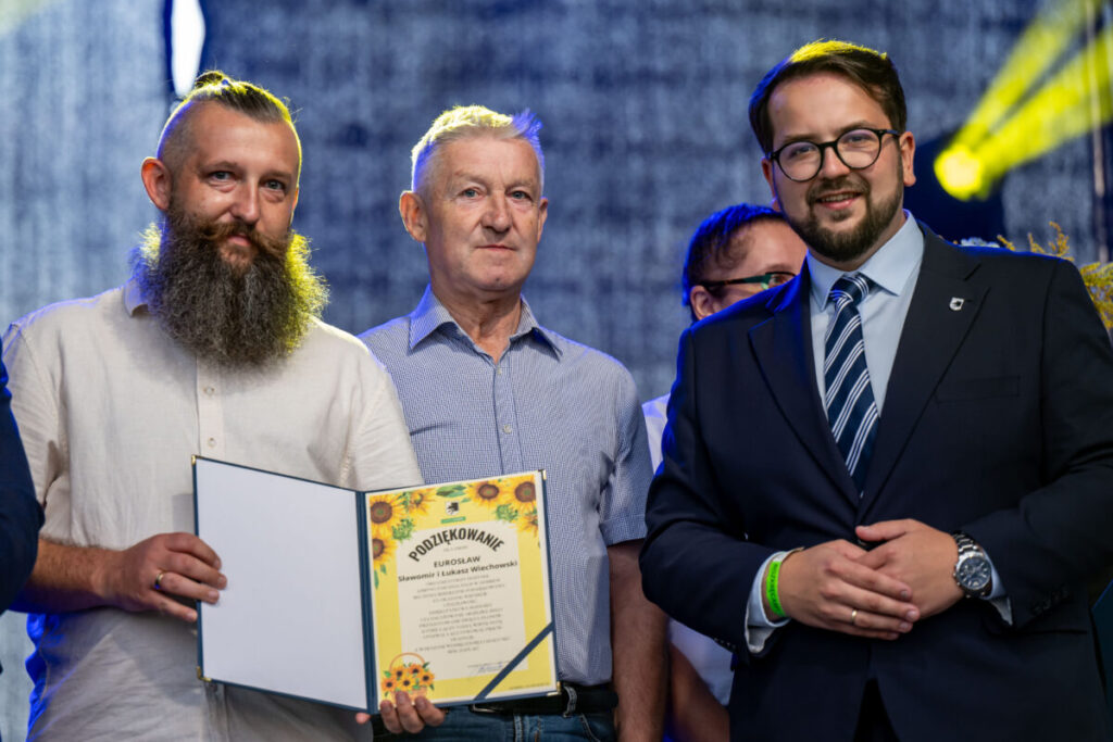 Na zdjęciu trzech mężczyzn. Jeden trzyma certyfikat z kolorowymi słonecznikami na żółtym tle. Tło jest ciemnoniebieskie z żółtym światłem.