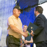 Nie wiem, kim są osoby na zdjęciu. Starszy mężczyzna w beżowej koszulce podaje dłoń mężczyźnie w garniturze z teczką. W tle są dwie osoby, tło jest ciemne z niebieskim odcieniem.