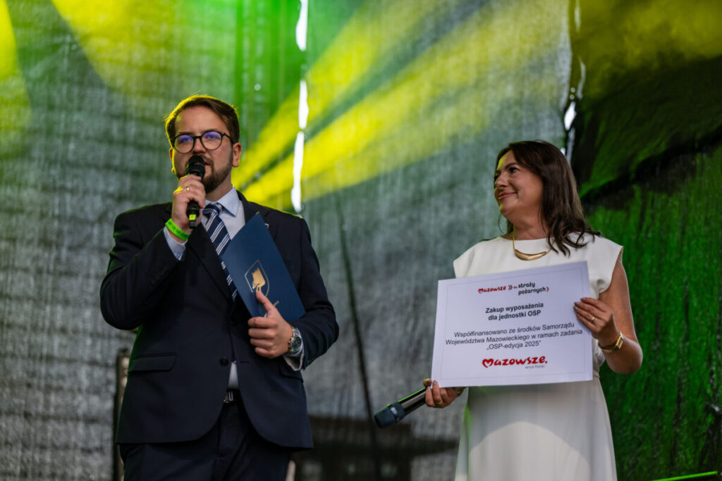 Dwóch ludzi na tle zielono-żółtego światła, mężczyzna w garniturze z mikrofonem, kobieta w bieli trzyma planszę.