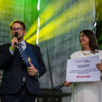 Dwóch ludzi na tle zielono-żółtego światła, mężczyzna w garniturze z mikrofonem, kobieta w bieli trzyma planszę.