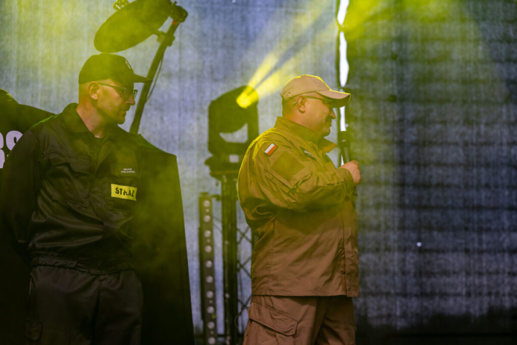 Przy drewnianym stole siedzą kobiety które robią dekoracje świąteczne z żywymi kwiatami w kolorze żółtym obol każdej leżą styropianowe jajka. W tle regały biblioteczne z książkami.