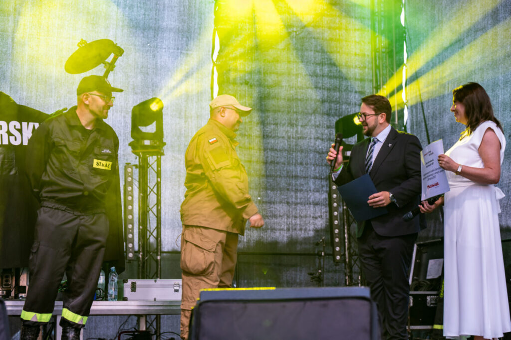 Na zdjęciu są cztery osoby na scenie. Tło jest ciemne z żółtym oświetleniem. Widać mundury i elegancki strój.
