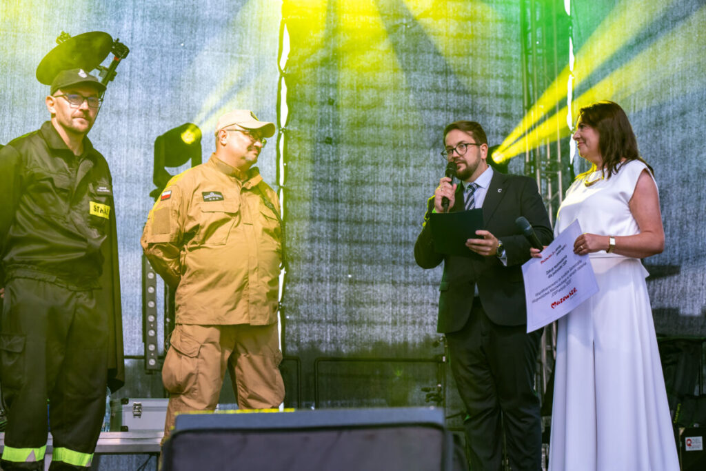 Na scenie cztery osoby: dwie w mundurach, jedna w garniturze z mikrofonem, kobieta w białej sukni trzyma certyfikat. Jasne reflektory na tle szarej kurtyny podkreślają postacie. Atmosfera oficjalna, oświetlenie w od