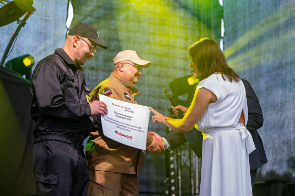 Na scenie są dwie osoby w mundurach, jedna trzyma tabliczkę. Kobieta w białej sukni podaje mikrofon. Tło jest zielone z reflektorami.