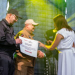 Na scenie są dwie osoby w mundurach, jedna trzyma tabliczkę. Kobieta w białej sukni podaje mikrofon. Tło jest zielone z reflektorami.