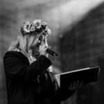 Kobieta z kwiatową wiankiem śpiewa do mikrofonu. Posiada otwartą książkę w dłoni. Czarnobiałe zdjęcie.