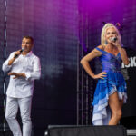 Na scenie występuje dwójka wokalistów z mikrofonami. Kobieta w niebieskiej sukience, mężczyzna w białym stroju. Tło ciemne z różowym oświetleniem.