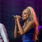 Na scenie kobieta w niebieskiej, błyszczącej sukni śpiewa do mikrofonu. Obok stoi mężczyzna w białej koszuli. Tło jest ciemne.