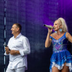 Dwójka artystów na scenie, mężczyzna w białej koszuli i kobieta w błyszczącej niebieskiej sukience. Jasno oświetlona scena. Ciemne tło.