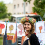 Osoba w kolorowym wianku stoi przed obrazami dziecięcych drzew na linkach. Tło to rozmyte budynki i zieleń. Uśmiechnięta kobieta.