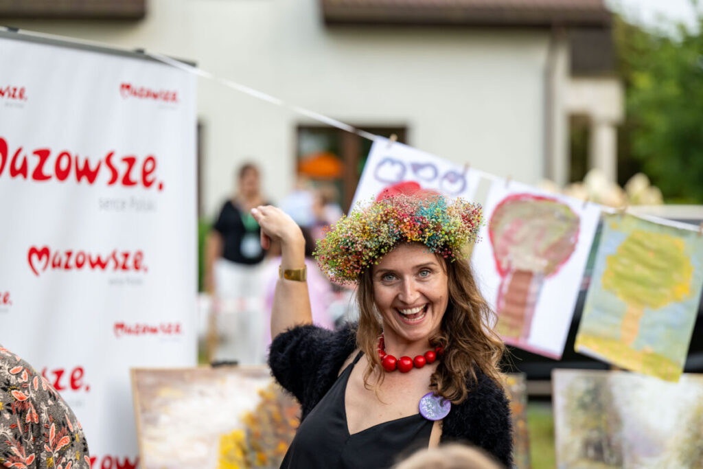 Na zdjęciu kobieta w kolorowym wianku, na tle banera Mazowsze. Ma na sobie czarną suknię i czerwony naszyjnik. W tle widoczne są dziecięce rysunki drzew.