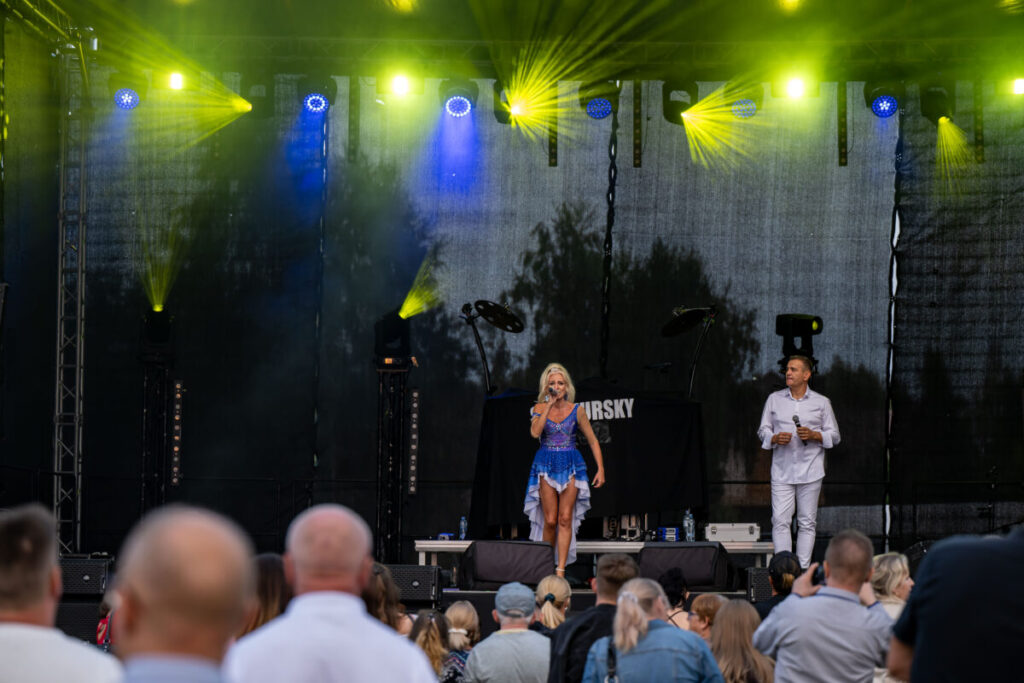 Na scenie kobieta w niebieskiej sukni śpiewa, obok stoi mężczyzna. Tło to czarne zasłony, żółte światła sceniczne. Publiczność widoczna od tyłu.