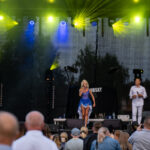 Na scenie kobieta w niebieskiej sukni śpiewa, obok stoi mężczyzna. Tło to czarne zasłony, żółte światła sceniczne. Publiczność widoczna od tyłu.