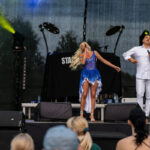 Dwie osoby śpiewają na scenie, kobieta w niebieskiej sukience, mężczyzna w białym stroju. Czarne tło z reflektorami.