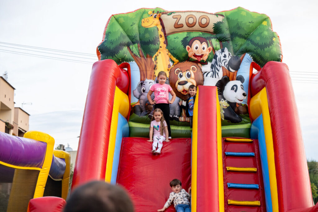 Dzieci zjeżdżają z kolorowej dmuchanej zjeżdżalni z motywem zoo. Tło pełne jest zieleni i uśmiechniętych zwierząt. Wokół jasne niebo.