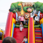 Dzieci zjeżdżają z kolorowej dmuchanej zjeżdżalni z motywem zoo. Tło pełne jest zieleni i uśmiechniętych zwierząt. Wokół jasne niebo.